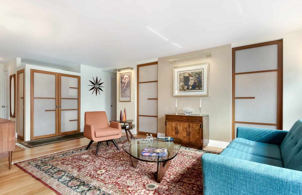 A stylish living room featuring a mix of mid-century modern and contemporary design elements. The room includes a peach-colored armchair, a glass coffee table with wooden legs, and a blue sofa, all arranged on a patterned red area rug. The walls are adorned with large, framed artwork, and the room is accented by sliding doors with frosted inserts framed in wood.