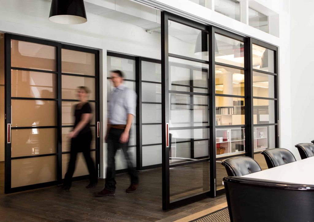 Two people walk through a hallway that is divided by a series of sleek, black-framed sliding doors.