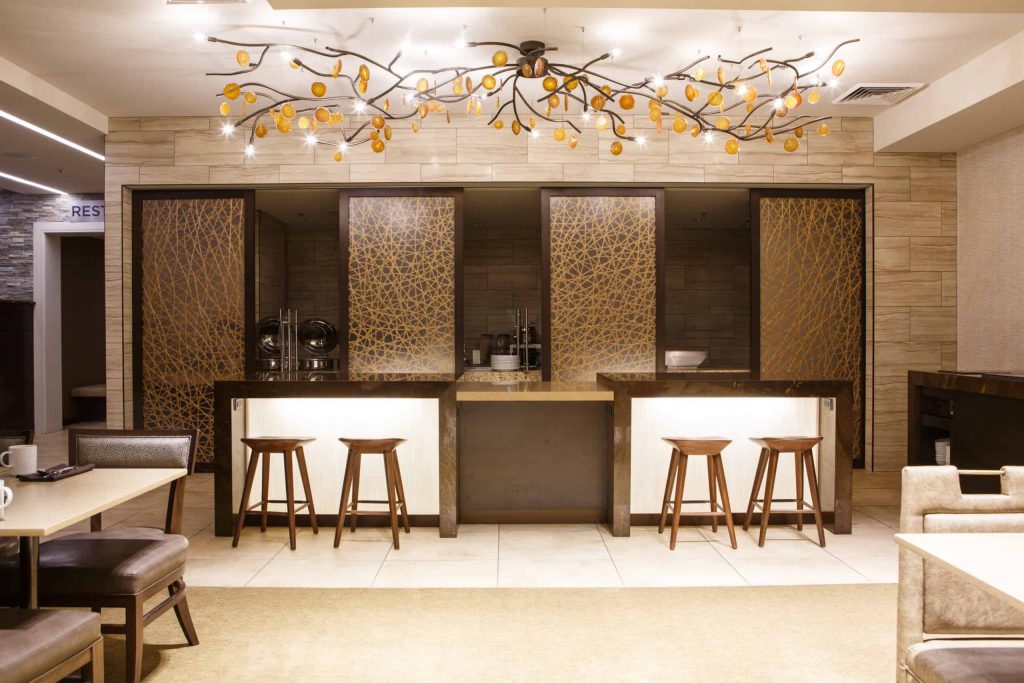 A modern bar area with high stools and a countertop, illuminated by a unique chandelier featuring a branch-like design with amber-colored accents. The background includes decorative panels with intricate patterns, creating a sophisticated and stylish environment.