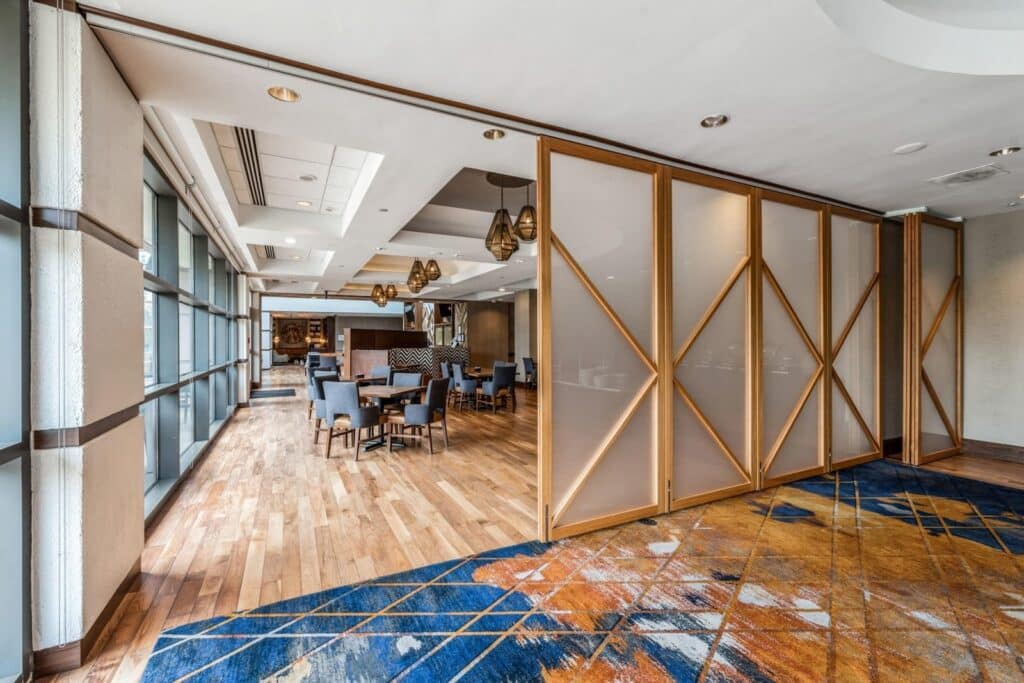 A stunning, cherry-wood sliding door system in the Sheraton Austin Hotel. The room divider is half-opened to reveal the common space past the doors. Acrylic panels allow ample light to flow between rooms while offering privacy, and the stacking system stows the divider neatly in the corner of the room.