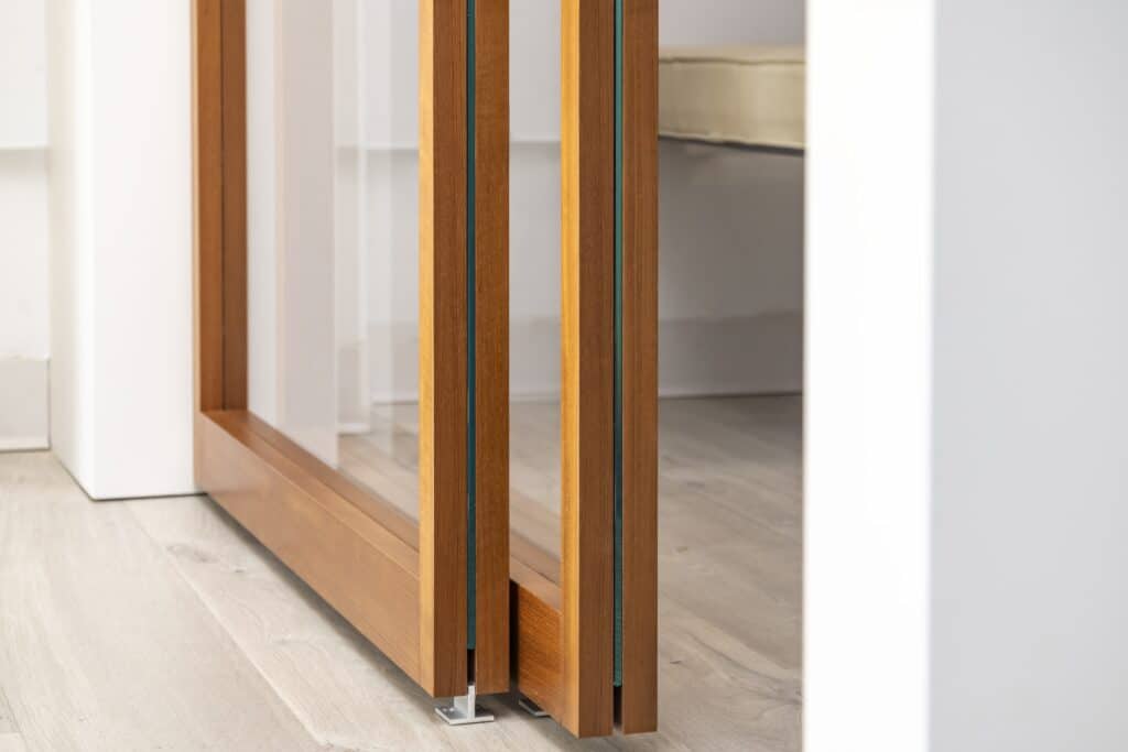 Close-up of the bottom of a wooden-framed glass sliding door, highlighting the craftsmanship of the frame and the smooth sliding mechanism. The door's glass panels reflect light, complementing the light-toned flooring in the modern interior.